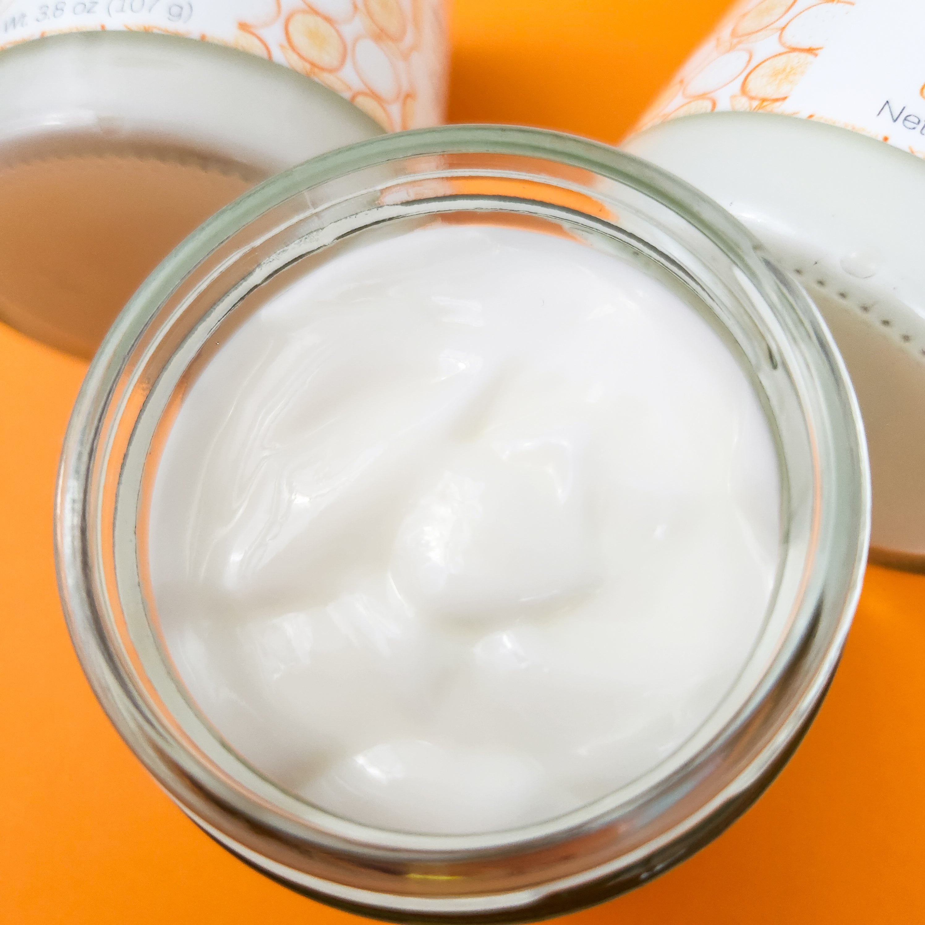 A closeup of an open jar of Citrus Bliss Body Lotion, showcasing the white lotion inside. Orange background.