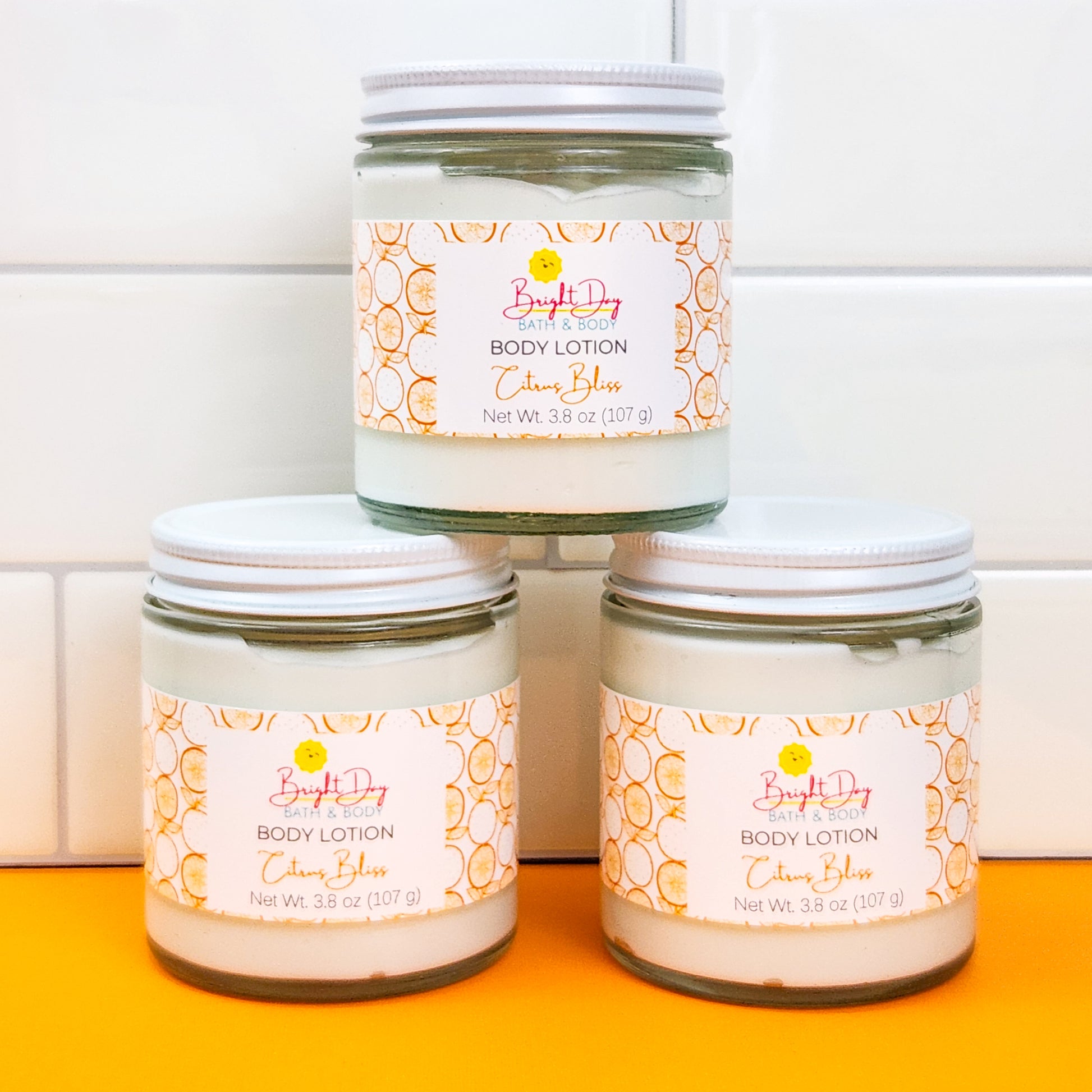 Three jars of Citrus Bliss Body Lotion stacked like a pyramid, on a white and orange background