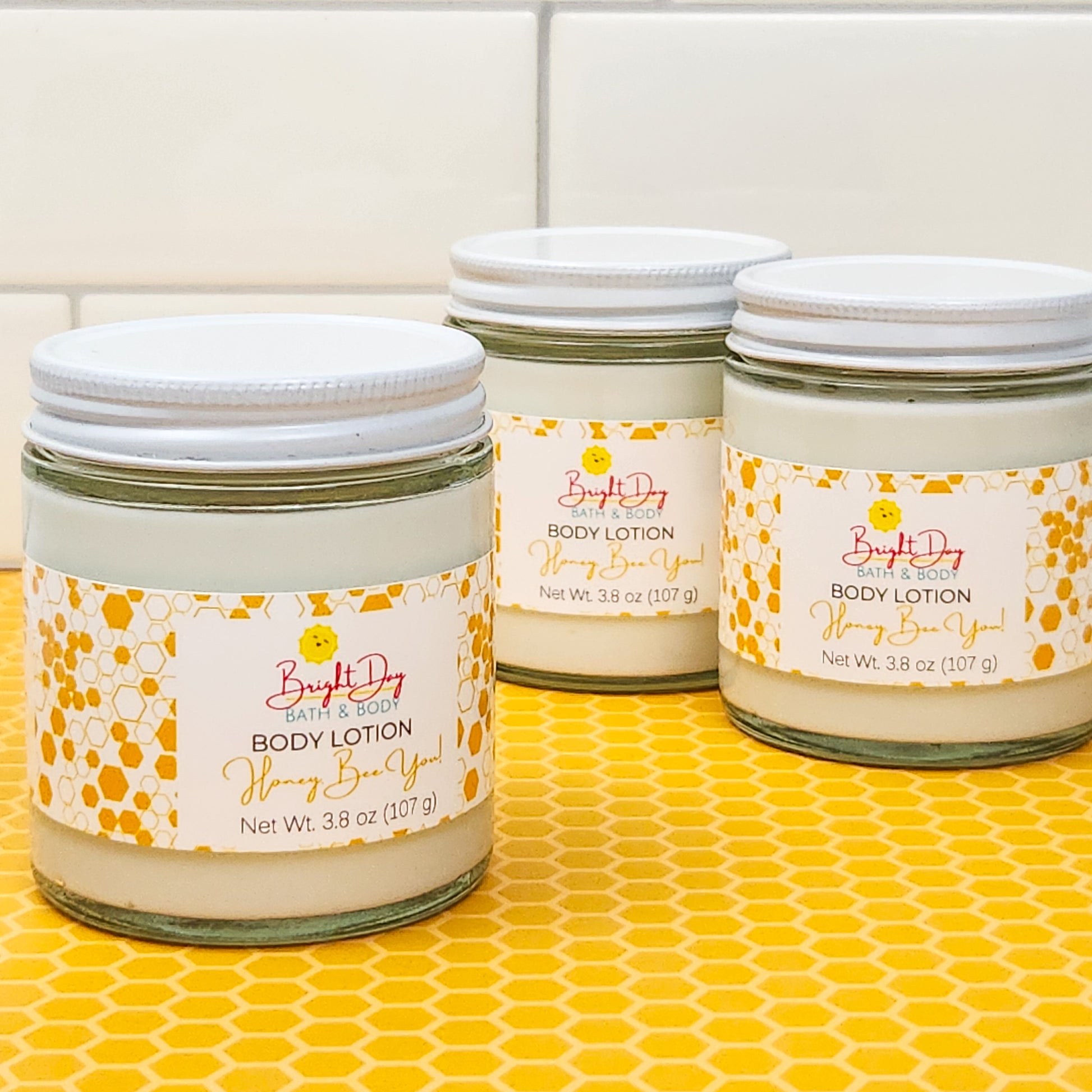 3 jars of Honey, Bee You Body Lotion, on a tan honeycomb background.