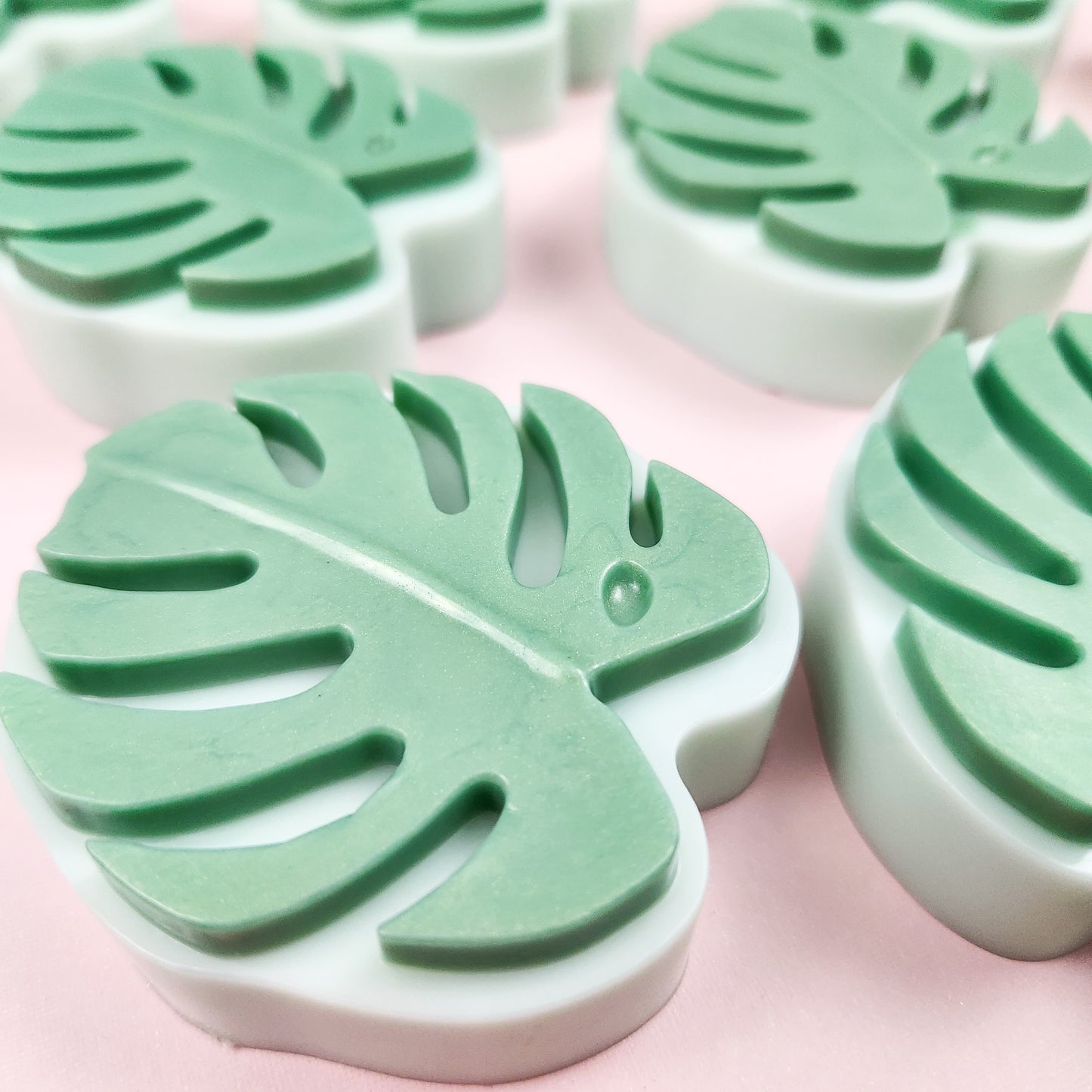 Close up of a light green soap in the shape of monstera leaf, on a pink background. Additional monsters leaf soaps in the background. 