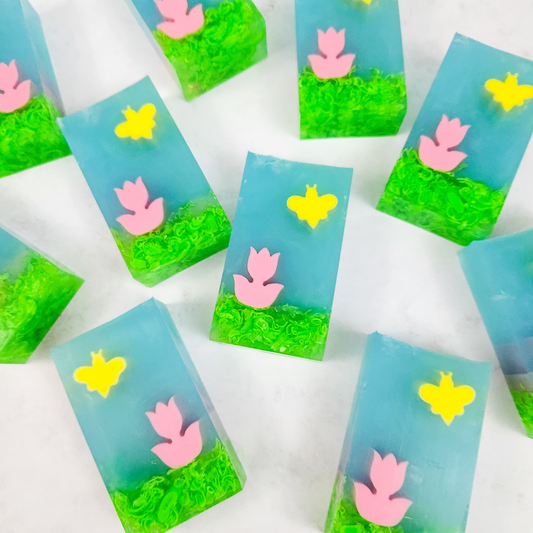 Soap bars in the design of a pink tulip on green grass, with a yellow bee in a blue sky.