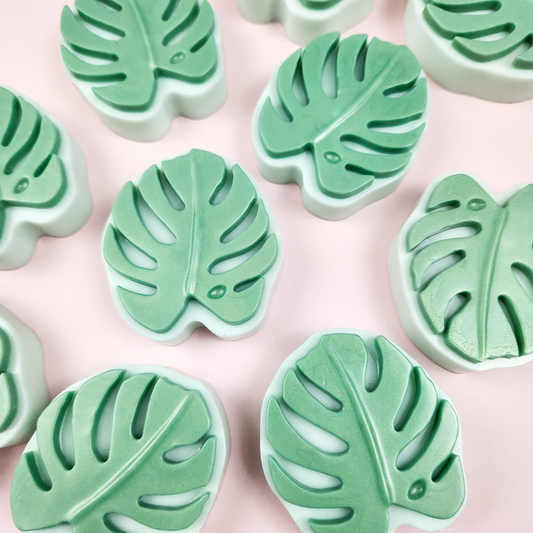 Several light green soaps in the shapes of monstera leaves, on a pink background.