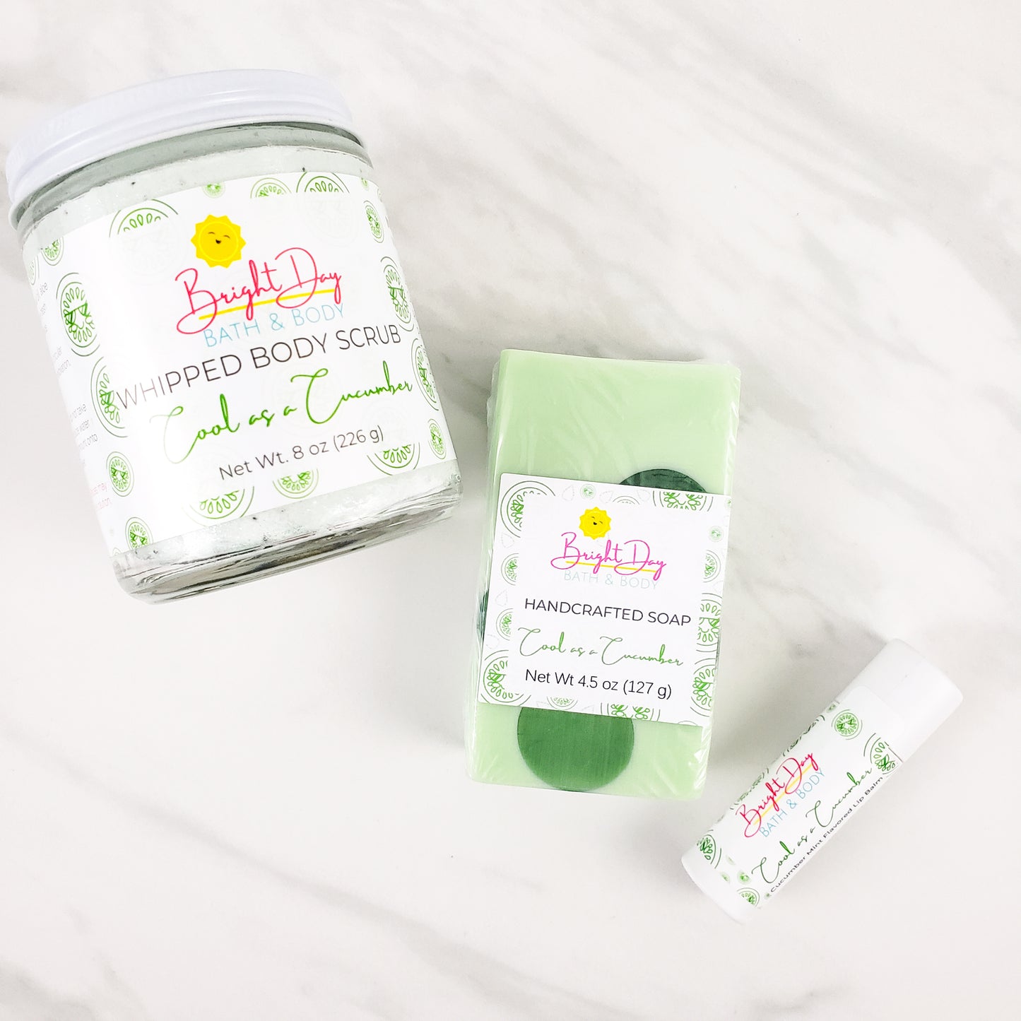 A Cool as a Cucumber body scrub, soap bar and lip balm on a marble background