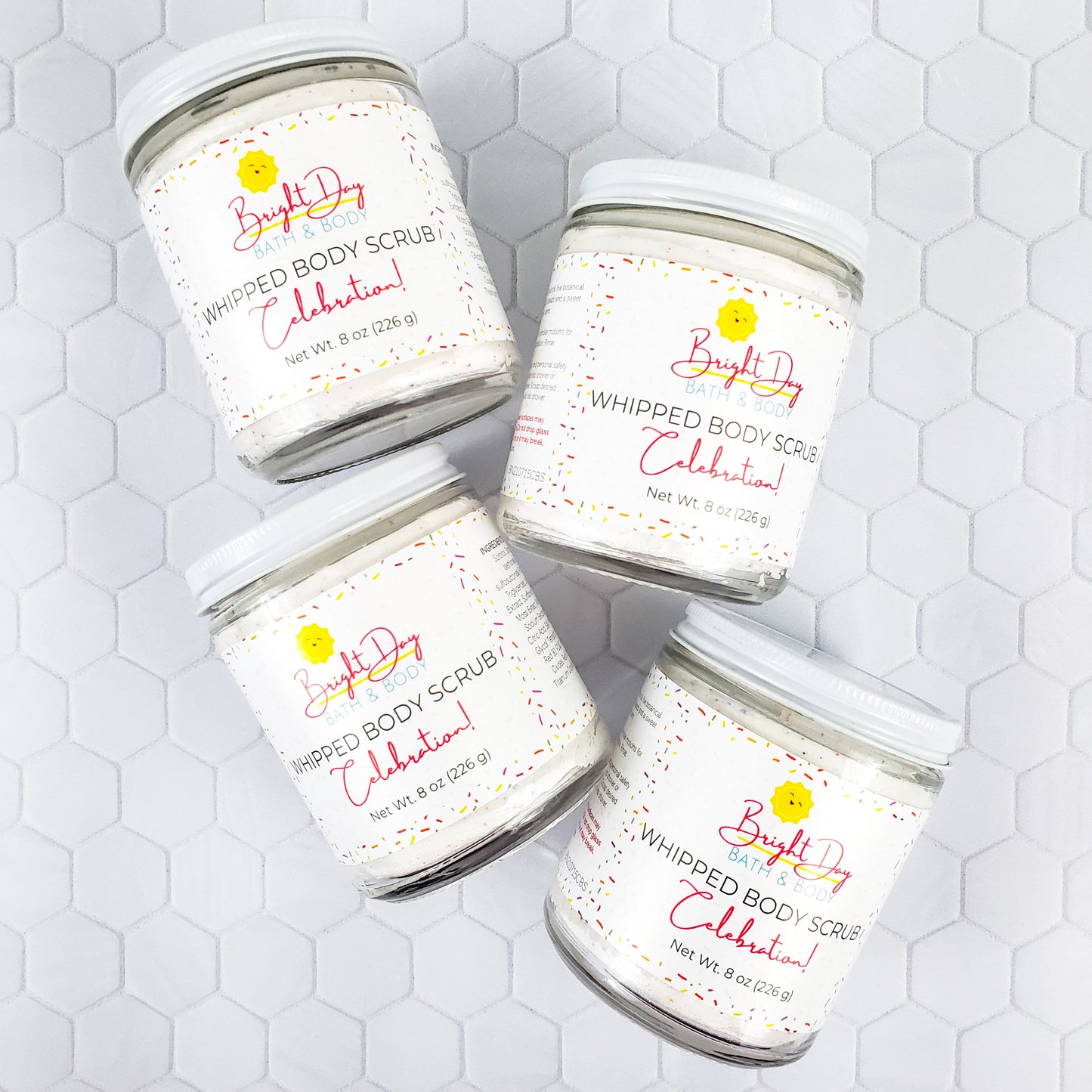 Four Celebration Body Scrub Jars on a tile background