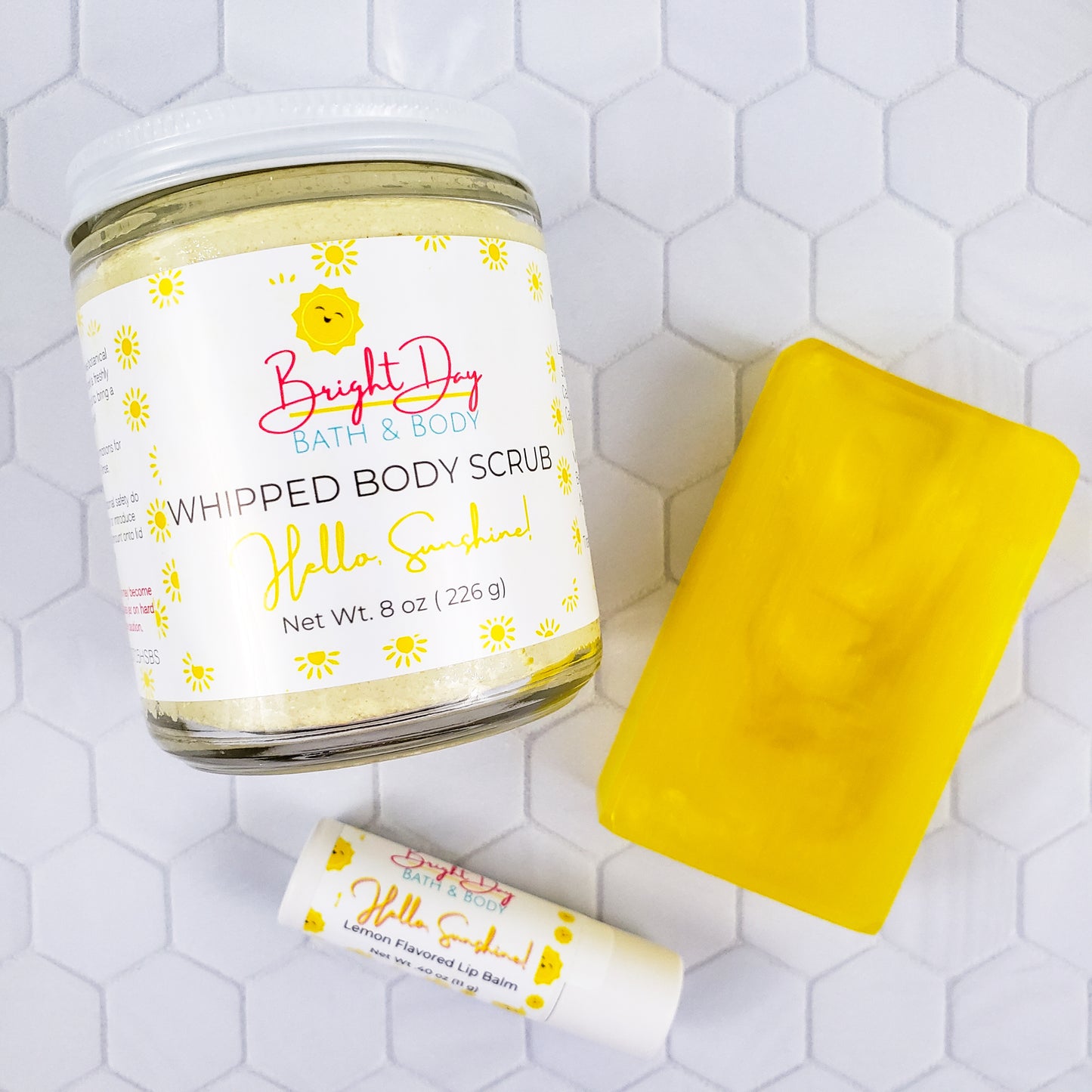 a Hello Sunshine body scrub, soap bar and lip balm on a tile background