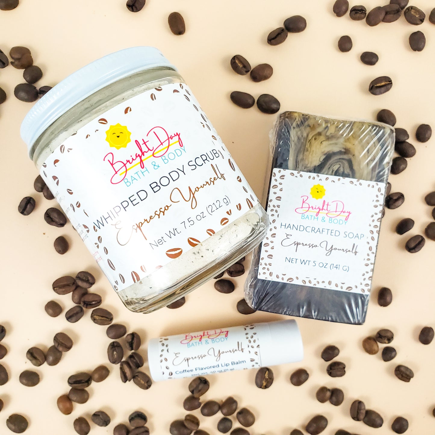 a Espresso Yourself body scrub, soap bar & lip balm on a tan background, surrounded by coffee beans