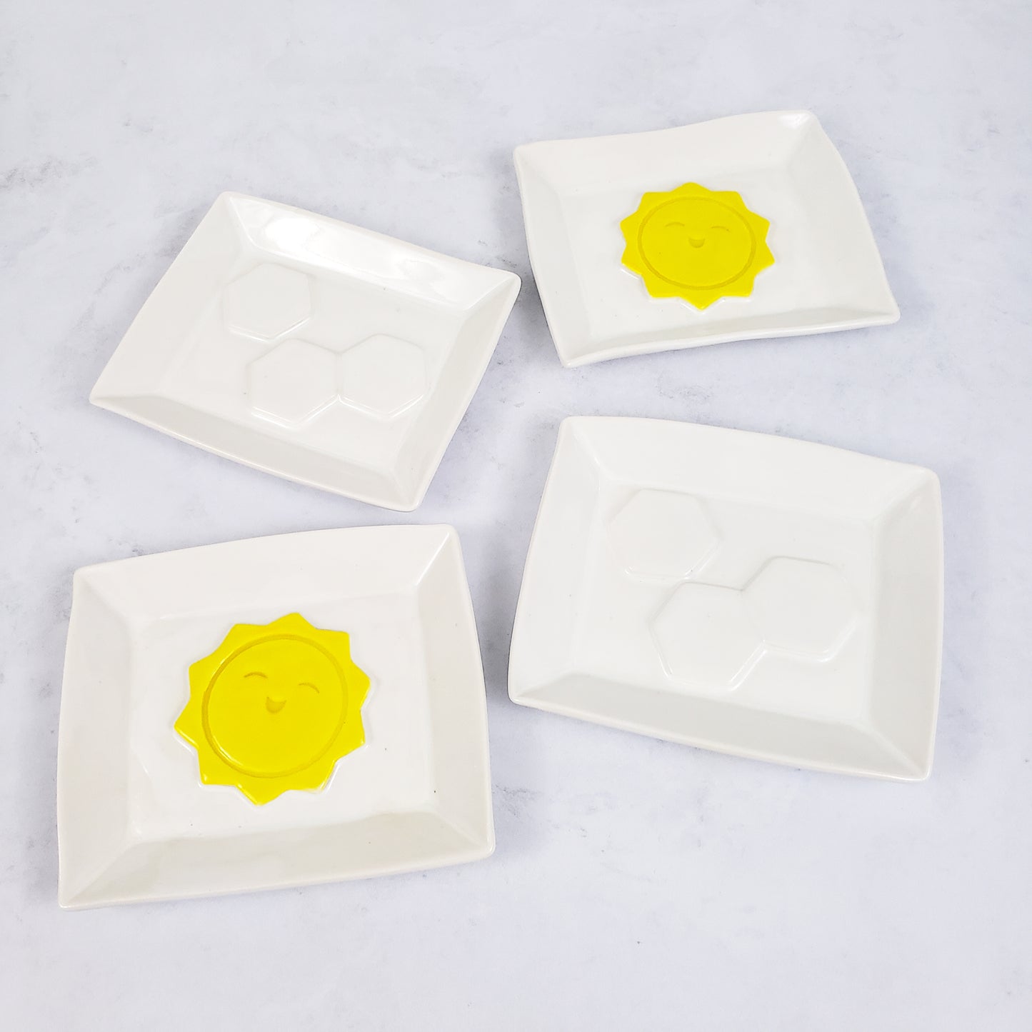 An assortment of white rectangular soap dishes, two with white hexagons in the middle,  two with sunshines in the middle