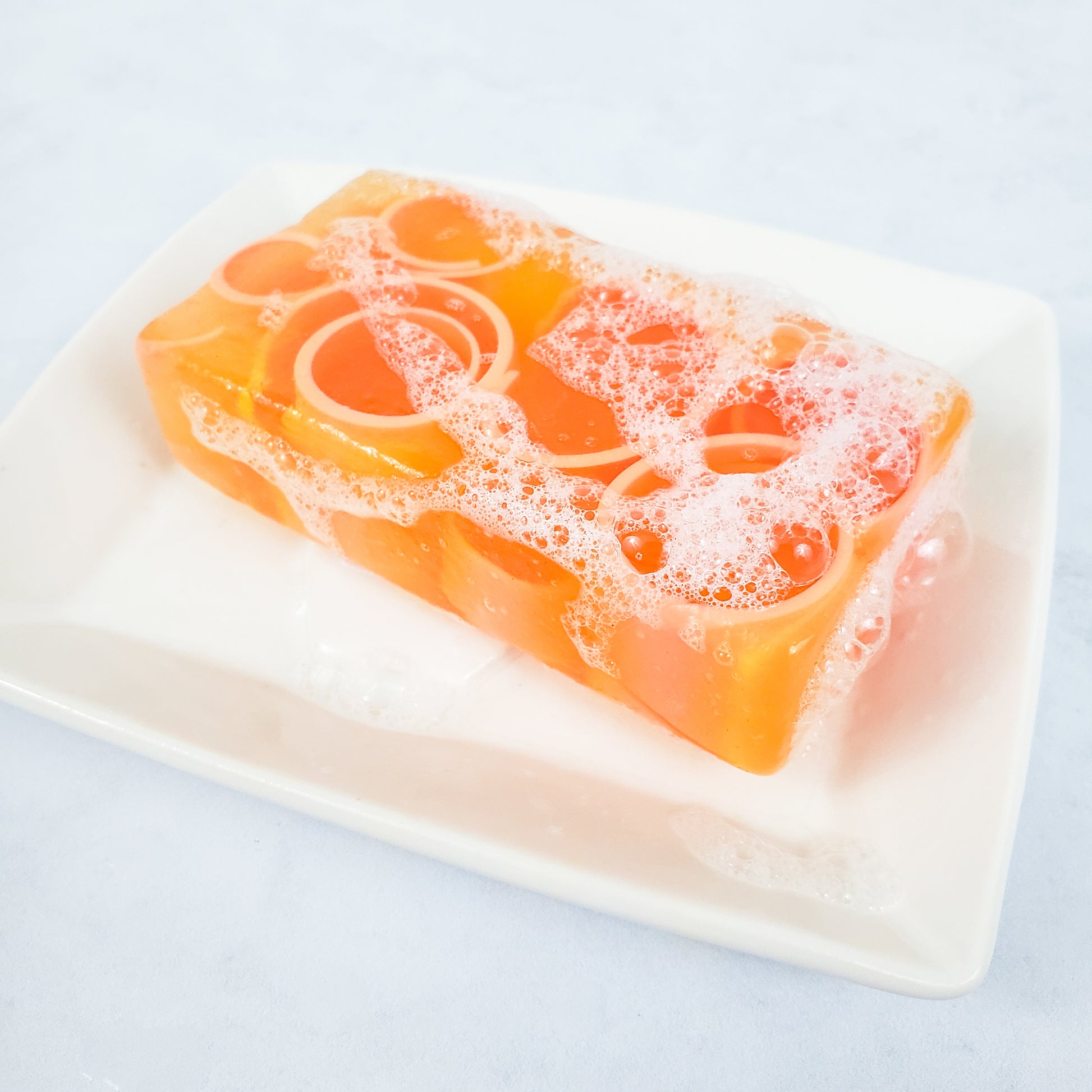 An orange bubbly soap on top of a white rectangle soap dish. On a white background.