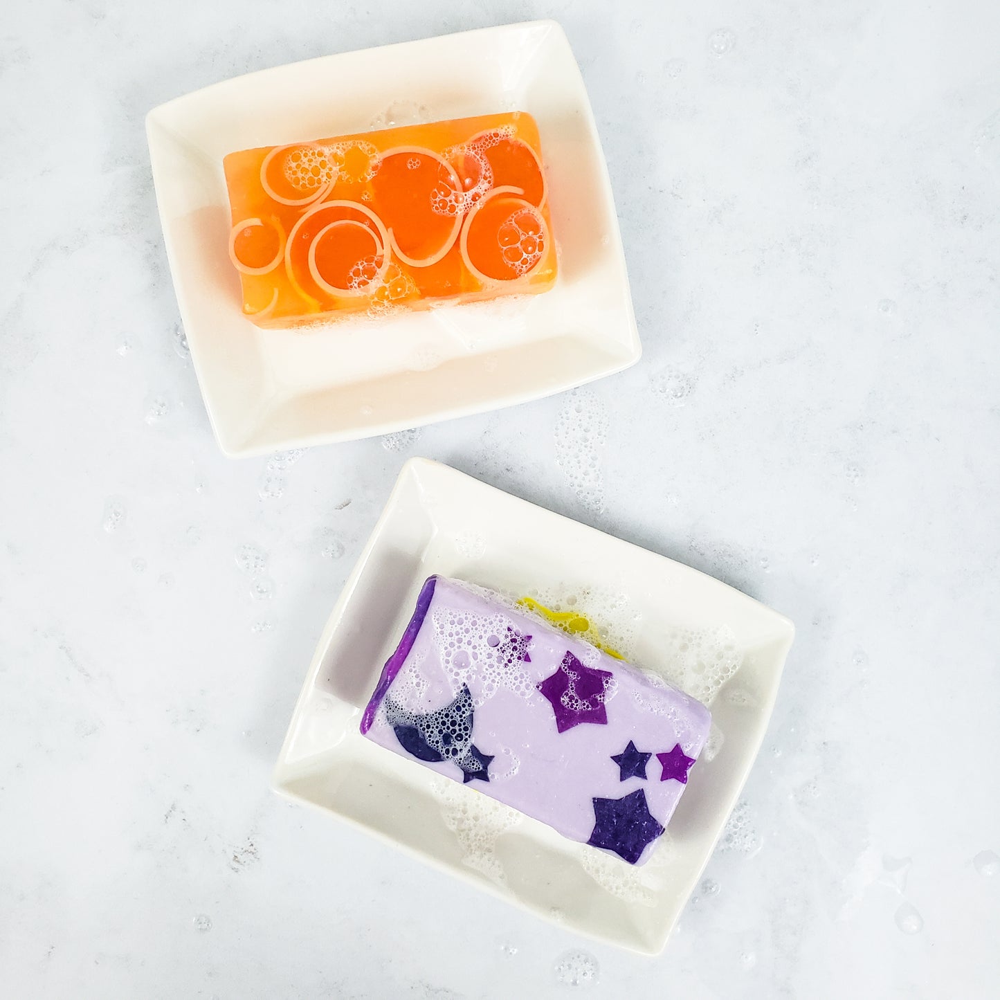 Two white rectangular soap dishes on a white background. One has an orange soap on top, one has a purple soap with stars on it.