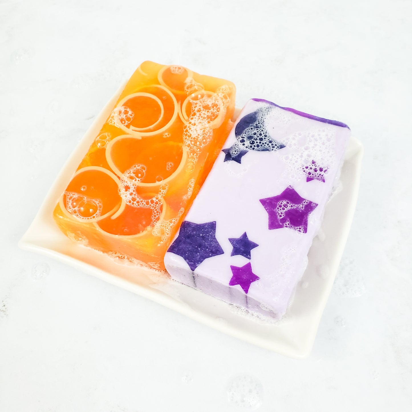 Two bars of soap (one orange, one purple with stars) on top of a white rectangle soap dish. On a white background.