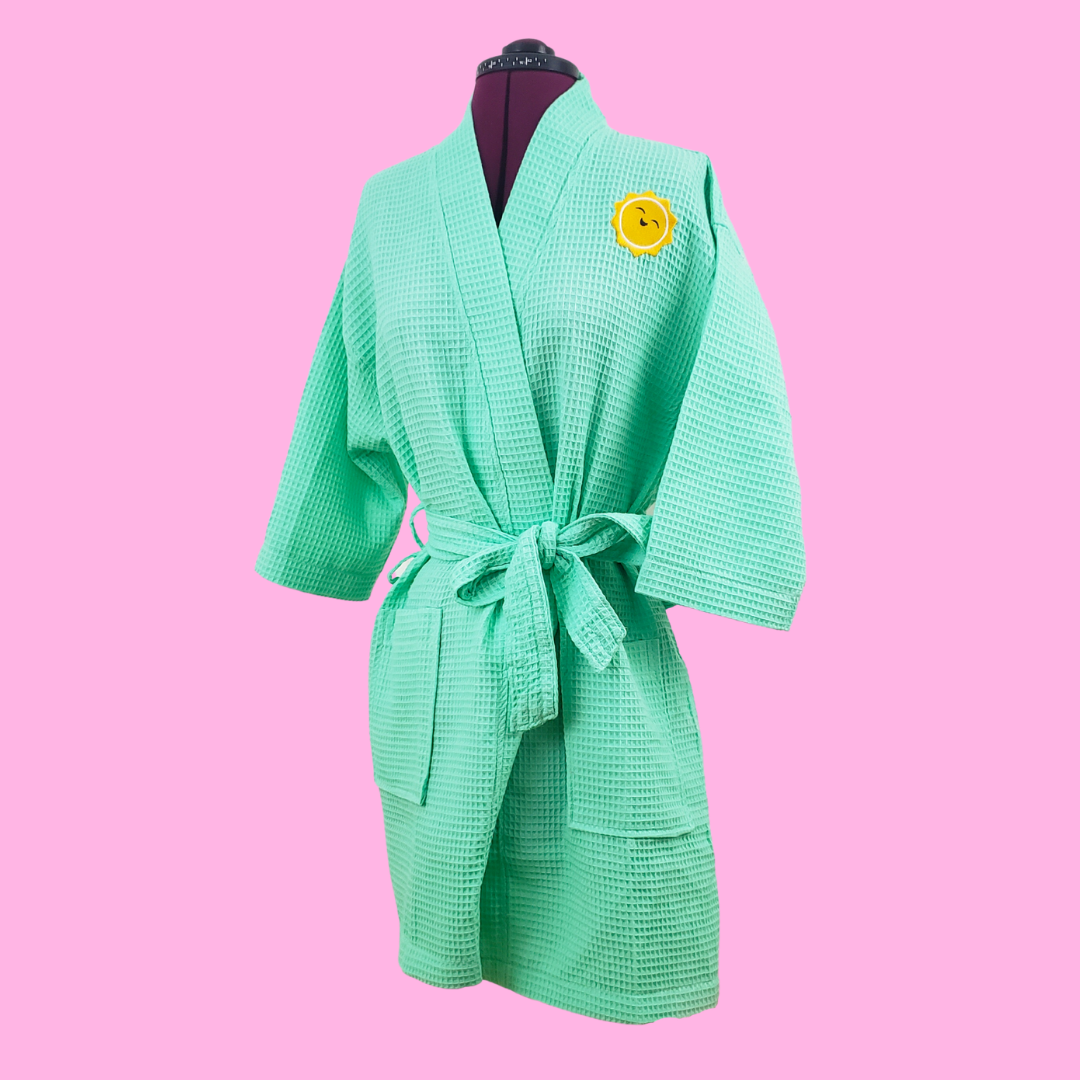 A sea green robe with a yellow sunshine on the top left shoulder. Photo has a pink background